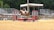 A horse and rider are in mid-jump over an obstacle that resembles a small brick wall on a sandy competition ground. In the background, there are empty bleachers and a canopy structure. The setting suggests an equestrian event, and the area is surrounded by lush green trees.