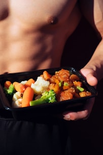 A shirtless person holds a black meal container filled with colorful vegetables and a savory dish, likely including broccoli, carrots, cauliflower, and a saucy protein mix.