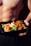 A shirtless person holds a black meal container filled with colorful vegetables and a savory dish, likely including broccoli, carrots, cauliflower, and a saucy protein mix.