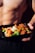 A shirtless person holds a black meal container filled with colorful vegetables and a savory dish, likely including broccoli, carrots, cauliflower, and a saucy protein mix.