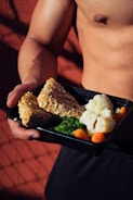 A person holding a meal in a black container featuring cooked cauliflower, broccoli, baby carrots, and slices of nut-crusted entrée. The background is a textured red surface with shadows, and the person appears shirtless, emphasizing a focus on fitness or healthy living.