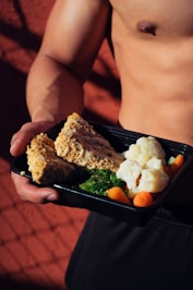 A person holding a meal in a black container featuring cooked cauliflower, broccoli, baby carrots, and slices of nut-crusted entrée. The background is a textured red surface with shadows, and the person appears shirtless, emphasizing a focus on fitness or healthy living.