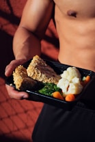 A person holding a meal in a black container featuring cooked cauliflower, broccoli, baby carrots, and slices of nut-crusted entrée. The background is a textured red surface with shadows, and the person appears shirtless, emphasizing a focus on fitness or healthy living.