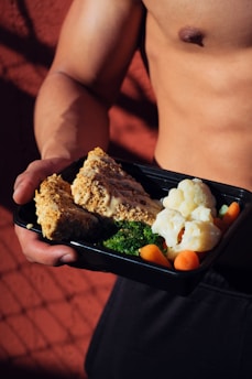 A person holding a meal in a black container featuring cooked cauliflower, broccoli, baby carrots, and slices of nut-crusted entrée. The background is a textured red surface with shadows, and the person appears shirtless, emphasizing a focus on fitness or healthy living.