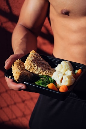A person holding a meal in a black container featuring cooked cauliflower, broccoli, baby carrots, and slices of nut-crusted entrée. The background is a textured red surface with shadows, and the person appears shirtless, emphasizing a focus on fitness or healthy living.