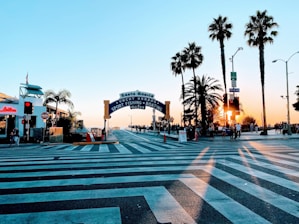 A crosswalk leads to a pier entrance with a sign reading 'Santa Monica Yacht Harbor Sport Fishing Boating Cafes.' Palm trees line the area, and the sun sets in the background, casting long shadows. People are walking around, and there's a traffic light indicating the pedestrian walkway.