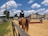 A person riding a horse is visible, wearing formal equestrian attire, including a helmet with a number. The scene is set in an outdoor equestrian arena, with obstacles for jumping. The area is surrounded by buildings and bleachers, and the sky is clear with scattered clouds.