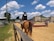 A person riding a horse is visible, wearing formal equestrian attire, including a helmet with a number. The scene is set in an outdoor equestrian arena, with obstacles for jumping. The area is surrounded by buildings and bleachers, and the sky is clear with scattered clouds.