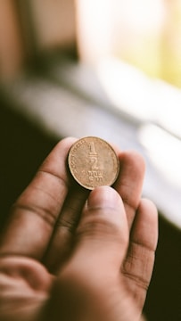 A hand is holding a coin with visible inscriptions in a slightly blurred background. The lighting creates a soft and warm ambiance, emphasizing the texture of the coin.