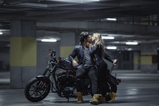 A man and a woman in black leather jackets are positioned on a motorcycle in a dimly lit parking garage. They are leaning towards each other in an intimate gesture, and the scene suggests a sense of adventure and romance. The lighting is low, creating a dramatic and moody atmosphere.
