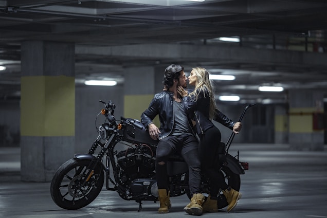 A man and a woman in black leather jackets are positioned on a motorcycle in a dimly lit parking garage. They are leaning towards each other in an intimate gesture, and the scene suggests a sense of adventure and romance. The lighting is low, creating a dramatic and moody atmosphere.