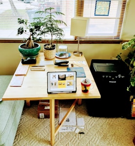 A wooden table is set in a cozy room with sunlight streaming through the blinds. On the table, there are two potted plants, a lamp, a tablet displaying various images, office supplies, and a coffee mug. A paper shredder is situated beside the table. The area has a calm and organized appearance.