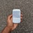 A hand holding a white smartphone with a QWERTY keyboard. The screen displays static or noise, echoing the gravel-like texture in the background.