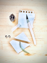 A small woodworking or crafting knife lays on a notepad with a few sheets torn out, set atop a wooden table. There are two metallic fasteners and a broken piece of paper with a pine needle placed across it. A decorative wooden object with intricate carvings is also present.