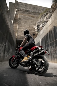 A motorcyclist wearing a black helmet and black clothing sits on a red and black motorcycle, positioned in an urban environment. The surrounding area consists of tall concrete walls with graffiti, and a large, rugged wall looms ahead, giving the impression of an underpass or alley.