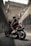 A motorcyclist wearing a black helmet and black clothing sits on a red and black motorcycle, positioned in an urban environment. The surrounding area consists of tall concrete walls with graffiti, and a large, rugged wall looms ahead, giving the impression of an underpass or alley.