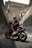 A motorcyclist wearing a black helmet and black clothing sits on a red and black motorcycle, positioned in an urban environment. The surrounding area consists of tall concrete walls with graffiti, and a large, rugged wall looms ahead, giving the impression of an underpass or alley.