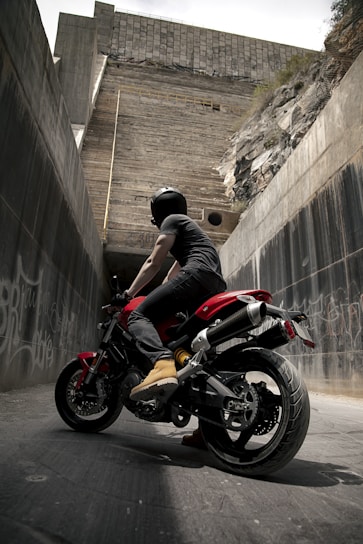A motorcyclist wearing a black helmet and black clothing sits on a red and black motorcycle, positioned in an urban environment. The surrounding area consists of tall concrete walls with graffiti, and a large, rugged wall looms ahead, giving the impression of an underpass or alley.