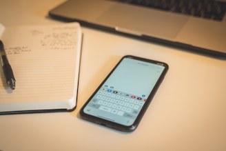 A smartphone displaying a keyboard is placed on a desk next to an open notebook with handwritten notes. A pen is positioned on the notebook, and a laptop is partially visible at the top of the image.