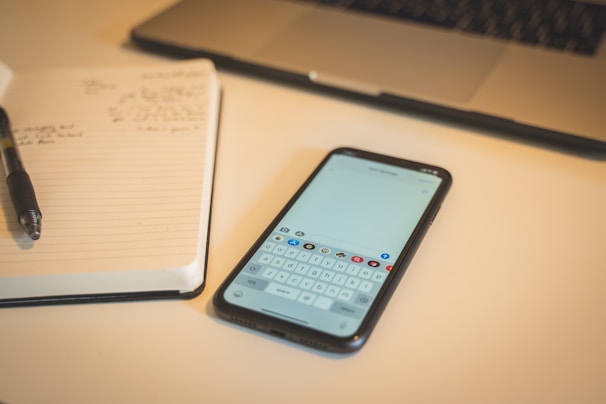 A smartphone displaying a keyboard is placed on a desk next to an open notebook with handwritten notes. A pen is positioned on the notebook, and a laptop is partially visible at the top of the image.