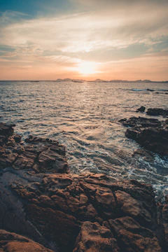 Sunset over a rocky coastline with waves gently crashing and a lighthouse in the distance.