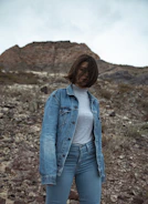 Model wearing a burnt rust jacket standing against a rocky mountain backdrop.