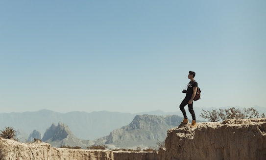 A person stands confidently on the edge of a rocky cliff, gazing out over a vast landscape of distant mountains under a clear blue sky. The individual is equipped with a backpack and a camera, suggesting they are exploring or photographing the scenery.