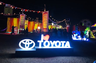A nighttime festival or fair setting with bright neon signs and colorful stalls. The illuminated sign prominently displays the Toyota logo and the words 'I love Toyota' at the forefront. Strung lights are overhead, creating a festive atmosphere. People are walking and gathering around various stalls that are decorated with vibrant colors like yellow and orange. Banners and flags accentuate the celebratory mood of the scene.