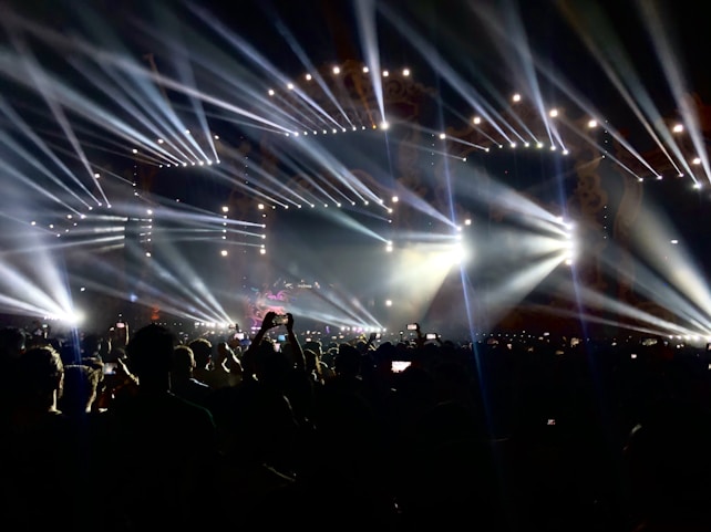 A crowded concert scene with a sea of people holding up phones to capture the moment. Bright, colorful stage lights shine in multiple directions, creating dramatic beams across the crowd. The atmosphere is lively and vibrant, with a large stage at the forefront.