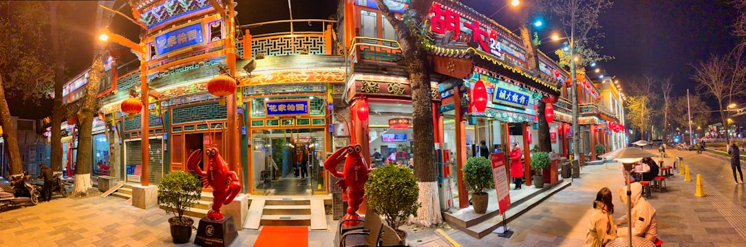 A vibrant street scene at night with a brightly lit building featuring traditional Chinese architectural elements such as ornate roof designs and red lanterns. Statues resembling red crayfish stand at the entrance. The area is bustling with activity, displaying signs with Chinese characters and illuminated signage.