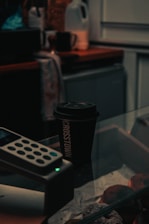 A dimly lit cafe scene features a black coffee cup with the word 'CROSSTOWN' printed vertically on it. The cup sits atop a glass display case filled with pastries. Nearby, the edge of a credit card machine is visible, and there is a slightly out-of-focus countertop with a jug and various items in the background.