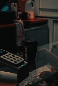 A dimly lit cafe scene features a black coffee cup with the word 'CROSSTOWN' printed vertically on it. The cup sits atop a glass display case filled with pastries. Nearby, the edge of a credit card machine is visible, and there is a slightly out-of-focus countertop with a jug and various items in the background.