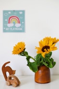 A small wooden toy car and an abstract wooden cat figurine sit on a white surface beside a brown vase containing vibrant sunflowers. A wall sign above features a colorful rainbow and the words 'My Playroom' surrounded by clouds.
