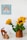 A small wooden toy car and an abstract wooden cat figurine sit on a white surface beside a brown vase containing vibrant sunflowers. A wall sign above features a colorful rainbow and the words 'My Playroom' surrounded by clouds.