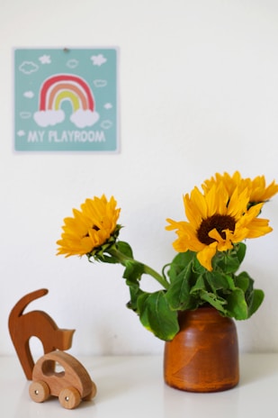 A small wooden toy car and an abstract wooden cat figurine sit on a white surface beside a brown vase containing vibrant sunflowers. A wall sign above features a colorful rainbow and the words 'My Playroom' surrounded by clouds.