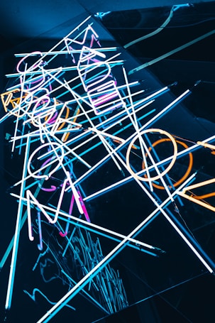 Neon light tubes arranged in an abstract pattern with overlapping lines and shapes, reflecting against a dark surface. The lights are in various colors including blue, orange, and pink, creating a vibrant and dynamic visual.