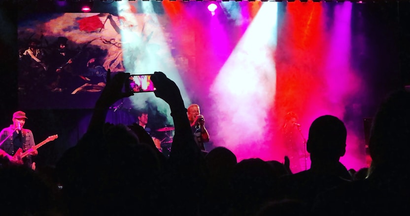 A live concert with vibrant stage lighting in pink, purple, and blue hues. A band performs on stage while a large screen displays artistic imagery in the background. The silhouettes of audience members holding up phones are visible in the foreground, capturing the performance.