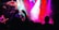A live concert with vibrant stage lighting in pink, purple, and blue hues. A band performs on stage while a large screen displays artistic imagery in the background. The silhouettes of audience members holding up phones are visible in the foreground, capturing the performance.