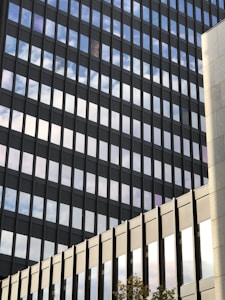 A modern office building with reflective glass windows displaying the sky. The geometric design features multiple horizontal and vertical lines creating a grid pattern. The facade is sleek and contemporary, showing reflections of clouds.