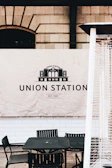 A historic-looking banner with the words 'Union Station Est. 1881' is displayed against a wall with a brick and block pattern. Below the banner, there is a set of outdoor black metal chairs and a table. Part of a tall patio heater with a metal grill is partially visible in the foreground.
