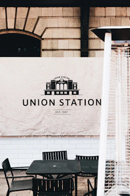 A historic-looking banner with the words 'Union Station Est. 1881' is displayed against a wall with a brick and block pattern. Below the banner, there is a set of outdoor black metal chairs and a table. Part of a tall patio heater with a metal grill is partially visible in the foreground.