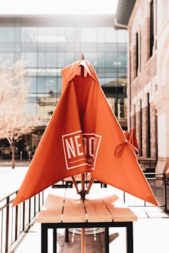 An outdoor seating area with a wooden table and an orange umbrella that has a logo on it. The umbrella is not opened fully and is positioned centrally on the table. The background features a modern glass building and trees with a hint of sunlight streaming in, creating a bright atmosphere.
