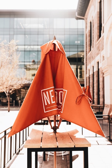 An outdoor seating area with a wooden table and an orange umbrella that has a logo on it. The umbrella is not opened fully and is positioned centrally on the table. The background features a modern glass building and trees with a hint of sunlight streaming in, creating a bright atmosphere.