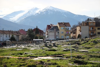 A scenic view of a small town with colorful houses in the foreground, nestled at the foot of a snow-capped mountain range. The landscape is dotted with scattered stone ruins, possibly an archaeological site, and green grass. Several people are walking around the area, suggesting an open, explorative environment. The sky is clear with bright daylight, highlighting the features of the town and surrounding nature.