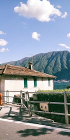 A serene landscape featuring a rustic house with a tiled roof positioned on a hillside. Behind the house, lush green mountains rise in the background, complemented by a clear blue sky with scattered fluffy clouds. The house has green shutters and sits adjacent to a paved road with a metal railing and a hedge.