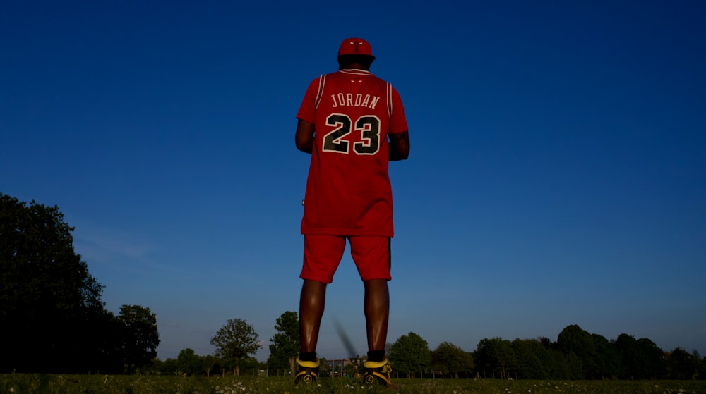 man in red and white jersey shirt and shorts standing on green grass field during daytime