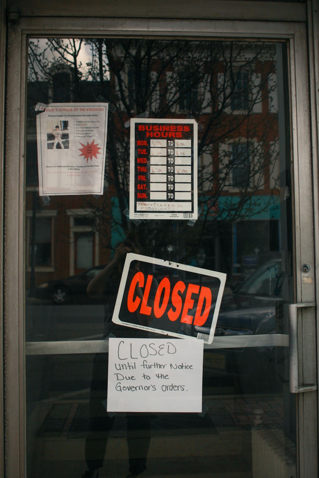 The glass door of a storefront displays several signs including a business hours chart with no times filled in, a large 'CLOSED' sign, and a handwritten note indicating closure until further notice due to the governor's orders. Reflections of the street and buildings across from the store can be seen on the glass.
