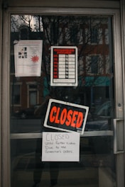 The glass door of a storefront displays several signs including a business hours chart with no times filled in, a large 'CLOSED' sign, and a handwritten note indicating closure until further notice due to the governor's orders. Reflections of the street and buildings across from the store can be seen on the glass.