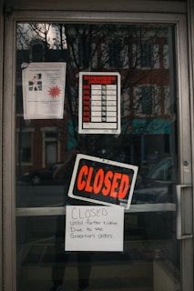 The glass door of a storefront displays several signs including a business hours chart with no times filled in, a large 'CLOSED' sign, and a handwritten note indicating closure until further notice due to the governor's orders. Reflections of the street and buildings across from the store can be seen on the glass.