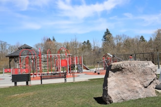 A playground with vibrant red and silver equipment, including climbing structures and slides, set within a fenced area. There is a large rock with an inscription marking the playpark. Surrounding the playground are trees and grass, and the sky is clear with a few clouds.